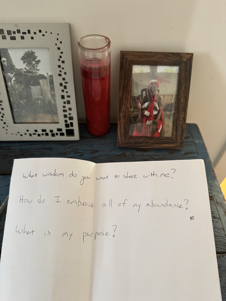 Open notebook with handwritten questions, 'What wisdom do you want to share with me?', 'How do I embrace all of my abundance?', 'What is my purpose?', beside a red candle and a framed photograph of a Bwiti elder.
